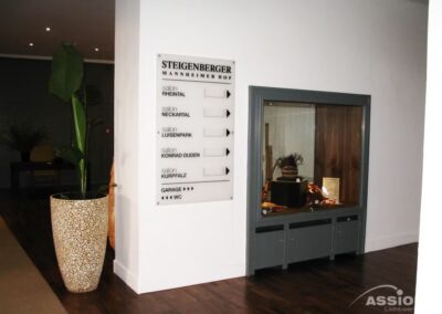 Ein Hotelflur mit einer Topfpflanze, einem Wegweiser mit Veranstaltungsräumen und einer Vitrine mit verschiedenen Gegenständen vor einer weißen Wand.