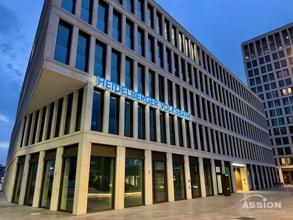 Leuchtschriften (1) XXX Modernes Bürogebäude mit großen Fenstern und beleuchteten Leuchtschriften auf einem blauen Schild mit der Aufschrift "Heidelberger Volksbank" an der Fassade, aufgenommen in der Abenddämmerung.