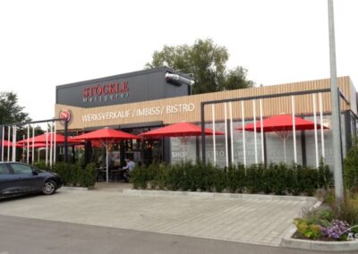 Moderne Schaufensterfront der Stöckle Metzgerei mit roten Sonnenschirmen, die die Sitzplätze im Freien beschatten, geparkten Autos davor und Schildern für Laden, Imbiss und Bistro.