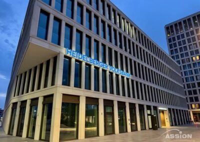 Modernes Bürogebäude mit großen Fenstern in der Abenddämmerung, mit beleuchtetem Schriftzug "Heidelberger Volksbank" an der Fassade.