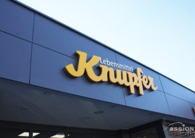 Schaufensterschild mit der Aufschrift "Lebensmittel Knupfer" in gelben und weißen Buchstaben auf einer modernen, dunklen Gebäudefassade unter klarem Himmel.