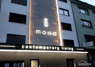 Eine moderne Gebäudefassade mit einem beleuchteten Schild mit der Aufschrift "mood" und einem Ananas-Symbol über den Worten "contemporary living" in weißen Buchstaben.