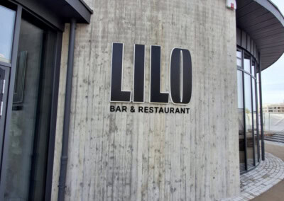 Außenansicht eines modernen geschwungenen Betongebäudes mit großem Schild "LILO BAR & RESTAURANT" an der Wand. Davor ein Weg aus Kopfsteinpflaster. Logo "ASSION" in der Ecke.