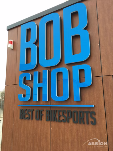 In großen blauen Buchstaben steht auf einer Holzwand "BOB SHOP", darunter in kleinerer Schrift "BEST OF BIKESPORTS". Über dem Schild ist eine kleine Sicherheitsvorrichtung angebracht.
