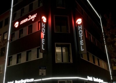 Ein modernes Hotelgebäude bei Nacht mit beleuchteten "Seven Days"- und "HOTEL"-Schildern an der Fassade; eine Person steht in der Nähe des Eingangs.
