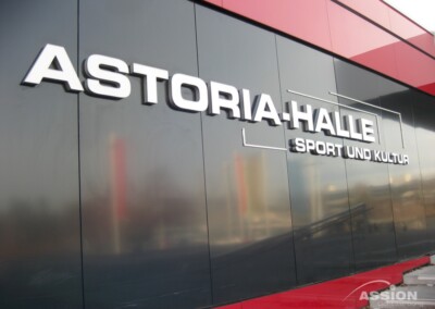 Eine große Außenbeschilderung zeigt "ASTORIA HALLE SPORT UND KULTUR" auf einer modernen Gebäudefassade mit reflektierenden Platten.