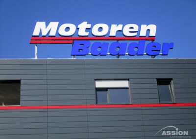Das Äußere eines grauen Gebäudes mit dem Schild "Motoren Baader" in großen Buchstaben auf dem Dach vor einem klaren blauen Himmel.