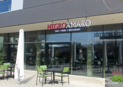 Sitzbereich im Freien vor dem Negroamaro Café, Music, Restaurant mit Tischen, Stühlen und einem geschlossenen Sonnenschirm an einem sonnigen Tag.