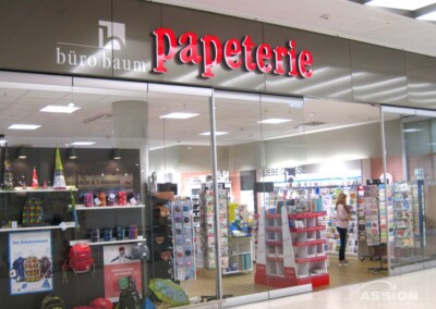 Schaufenster eines Schreibwarengeschäfts mit dem Namen "Papeterie bürobaum", das durch große Glasfenster in einem Einkaufszentrum Rucksäcke, Büroartikel und Grußkarten ausstellt.