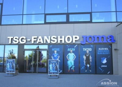 Außenansicht des TSG-Fanshops mit großem Joma-Schild und Werbepostern mit Fußballspielern in verschiedenen Trikots an den Fenstern.