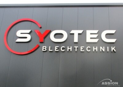 Firmenschild mit der Aufschrift "SYOTEC BLECHTECHNIK" in fetten Buchstaben auf einer grauen Wand, wobei der erste Buchstabe teilweise rot umrandet ist.