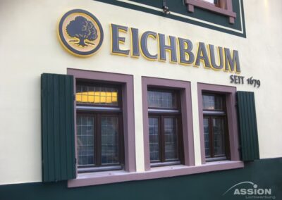 Das Äußere eines Gebäudes zeigt das Eichbaum-Logo, ein Baumsymbol und den Text "Eichbaum Seit 1679" über drei Fenstern mit dunkelgrünen Fensterläden.
