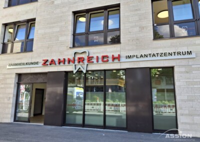Außenansicht einer Zahnklinik mit dem Namen ZAHNREICH IMPLANTATZENTRUM, mit großen Fenstern und beiger Steinfassade. Auf dem Schild der Klinik ist ein Zahnsymbol abgebildet.