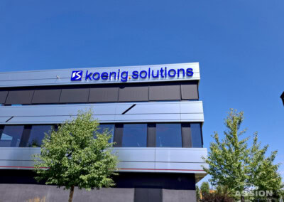 Ein modernes Gebäude mit einem "koenig.solutions"-Schild an der Außenseite, blauer Himmel über dem Kopf und Bäume im Vordergrund.