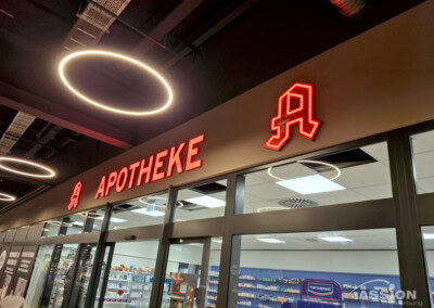 Ein Schaufenster mit dem roten Neonschild "APOTHEKE" über dem Eingang einer Apotheke; im Inneren sind Regale mit Produkten zu sehen.