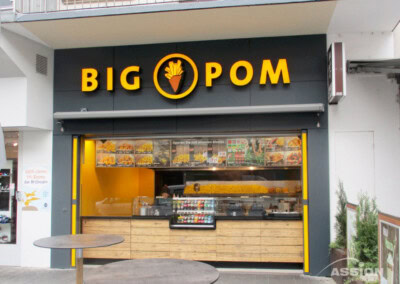 Die Schaufensterfront von BIG POM" ist mit einem gelben Schild und einer Theke mit Lebensmitteln versehen, im Inneren befinden sich Menütafeln und ein Grill.