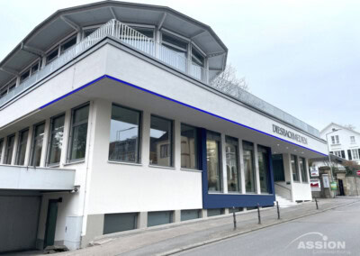 Modernes zweistöckiges weißes Gebäude mit großen Fenstern und einem Schild mit der Aufschrift "DIEBRAUCHICH.DE" an der oberen Fassade, an einer abschüssigen Straße gelegen.