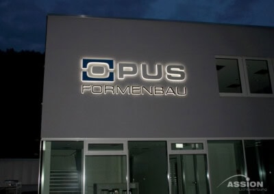 Das Äußere eines modernen Gebäudes bei Nacht mit der Leuchtreklame "OPUS FORMENBAU" über großen Fenstern und Glastüren.