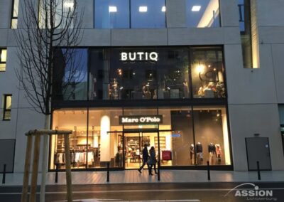 Schaufenster mit "BUTIQ" im Obergeschoss und "Marc O'Polo" im Erdgeschoss, mit zwei Personen, die auf einer nassen Straße vorbeigehen.