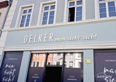 Schaufenster von DELKER Augenoptik mit dem Slogan "man sieht sich!" und der Nummer 30 auf dem Gebäude, mit großen Schaufenstern, in denen sich die umliegenden Gebäude spiegeln.