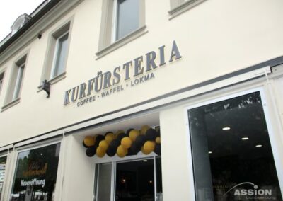 Das Äußere eines Cafés namens Kurfürsteria, mit einem Schild "Kaffee - Waffel - Lokma" über großen Fenstern und einem mit schwarzen und gelben Luftballons geschmückten Eingang.
