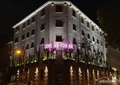Ein vierstöckiges Gebäude bei Nacht mit beleuchteten Fenstern und einer Leuchtreklame mit der Aufschrift "COME AS YOU ARE" an der Fassade. Auf der Straße darunter sind Autos geparkt.