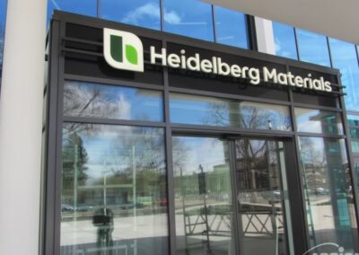 Gläserner Eingang eines Gebäudes mit beleuchteten Leuchtschriften, die das Logo und den Namen von Heidelberg Materials über der Tür anzeigen; im Glas sind Spiegelungen von Bäumen und Himmel sichtbar.