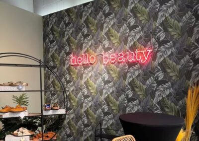 Eine Leuchtreklame mit der Aufschrift "Hallo Schönheit" ist an einer blattgemusterten Wand angebracht, daneben stehen ein Tisch, Stühle und ein Regal mit Gebäck.