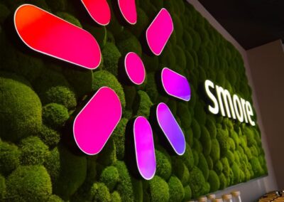 Eine Wand mit einem leuchtend rosafarbenen und lilafarbenen blumenähnlichen Logo und dem Wort "Smore" auf grünem Moosgrund über einer Theke mit Glasflaschen.
