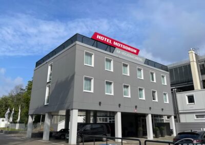 Ein modernes graues Gebäude mit der Aufschrift "Hotel Motodrom" zeigt eine auffällige Leuchttransparente, mehrere Fenster, darunter geparkte Autos und einen klaren Himmel im Hintergrund.