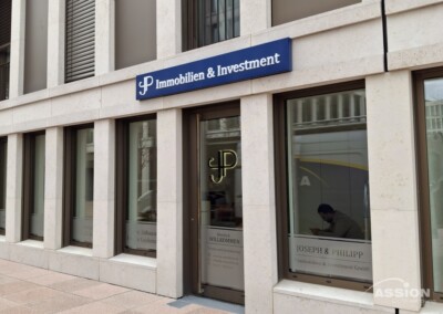 Straßenansicht eines Gebäudes mit der Aufschrift "Immobilien & Investment" über Glastüren und Fenstern, Innenansicht mit einer Person, die an einem Schreibtisch arbeitet.