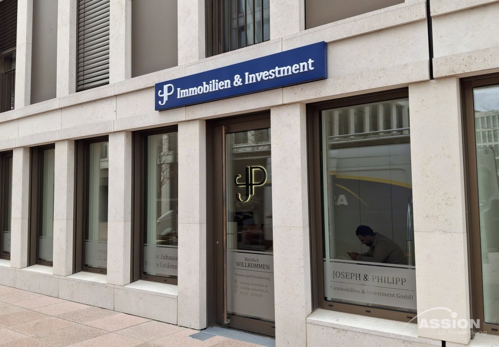 Straßenansicht des Büros von Joseph & Philipp Immobilien & Investment, mit großen Fenstern, einem Leuchttransparente-Schild darüber und einer Person, die im Inneren arbeitet.