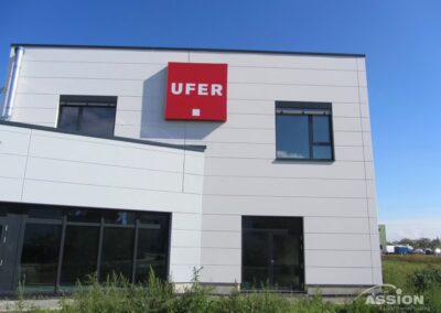 Modernes weißes Geschäftsgebäude mit einem roten Schild mit der Aufschrift "UFER" an der oberen Wand, unter einem klaren blauen Himmel.