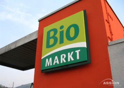 Ein rechteckiges Schild mit der Aufschrift "Bio MARKT" ist an einer orangefarbenen Wand außerhalb eines Gebäudes angebracht, wobei ein Teil der Dachkonstruktion sichtbar ist.