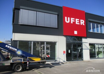 Ein modernes Geschäftsgebäude mit großen Fenstern und einem roten Schild mit der Aufschrift "UFER"; ein Fahrzeug mit einer Hebebühne mit der Aufschrift "ASSION" ist davor geparkt.