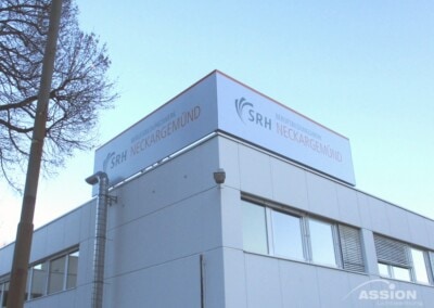 Ein modernes weißes Gebäude mit einem großen Schild auf dem Dach mit der Aufschrift "SRH Berufsbildungswerk Neckargemünd" vor einem klaren blauen Himmel.