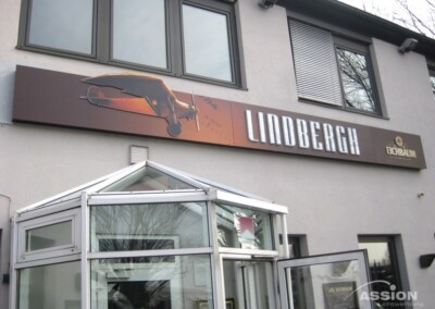 Eine Gebäudeaußenseite mit einem Schild, auf dem ein Flugzeug abgebildet ist und das Wort "LINDBERGH" in großen Buchstaben steht. Unter dem Schild sind gläserne Eingangstüren zu sehen.