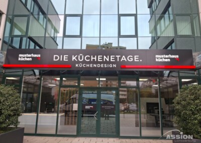 Schaufenster aus Glas mit einem schwarzen Schild mit der Aufschrift "DIE KÜCHENETAGE. KÜCHENDESIGN" und "musterhaus küchen"-Logos; im Hintergrund ein modernes Bürogebäude.