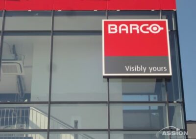 Auf einem rot-weißen Schild an einem Glasgebäude steht "BARCO Visibly yours".