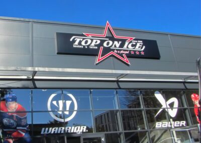 Außenansicht des Eishockey- und Sportgeschäfts "Top On Ice" mit großem Schild, blauem Himmel und Schaufenstergrafiken mit Eishockeyspielern und Sportmarkenlogos.