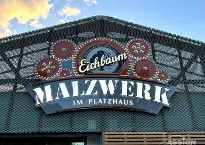 Ein Schild mit der Aufschrift "Eichbaum Malzwerk im Platzhaus" mit dekorativen Zahnrädern ist an einer Gebäudefassade aus Metallgitter angebracht.