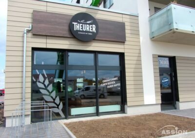 Außenansicht einer Bäckerei namens "Theurer" mit großen Fenstern, Schildern und einem kahlen Fleck Erde vor dem Eingang.