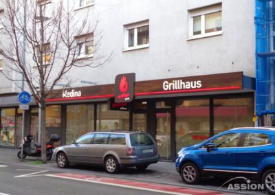 Straßenansicht eines Grillhaus-Restaurants mit einem braun-roten Schild, geparkten Autos davor und einem blattlosen Baum auf dem Gehweg.