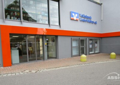 Außenansicht einer Volksbank-Filiale mit Glastüren, roten Akzenten und einem gepflasterten Gehweg; zwei gelbe Poller befinden sich vor dem Eingang.