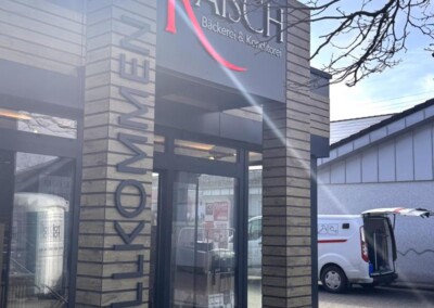 Eingang der Bäckerei mit dem Schild "RAISCH Bäckerei & Konditorei" über der Tür und der Aufschrift "WILLKOMMEN", die senkrecht an der Wand angebracht ist. Draußen ist ein Lieferwagen geparkt.