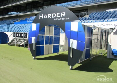 Ein blau-weißer Spielertunnel mit dem Schriftzug "Harder & Partner" auf dem Bogen, der sich am Rande eines Stadionfeldes mit blauen Sitzen im Hintergrund befindet.