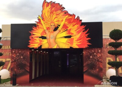 Der Eingang ist mit dem Bild eines Showgirls in einem gelben Federkostüm geschmückt, mit leuchtenden Federrequisiten über einem tunnelartigen Eingang, umgeben von Formschnittpflanzen.