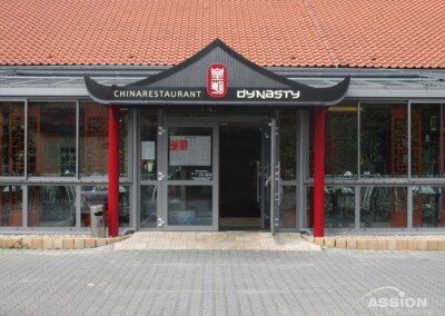 Vordereingang des chinesischen Restaurants Dynasty mit Glastüren, roten Säulen und einem Schild mit dem Namen des Restaurants darüber.