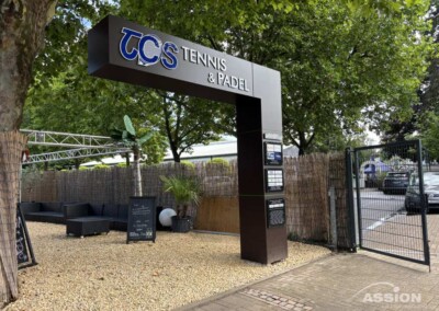 Eingang zu einem Tennis- und Padelclub, gekennzeichnet durch ein großes Schild mit der Aufschrift "TCS Tennis & Padel", mit Sitzgelegenheiten, Zaun und Bäumen im Hintergrund.