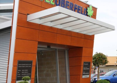 Moderner Gebäudeeingang mit orangefarbener Vertäfelung, weißem Vordach und dem Schriftzug "HOLZ OBERFELD" über den Türen; kleine Grünanlage im Vordergrund.